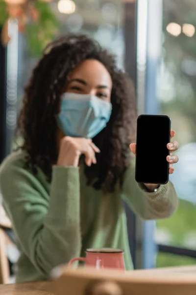 Smartphone avec écran blanc en main de la femme afro-américaine en masque médical sur fond flou — Photo de stock
