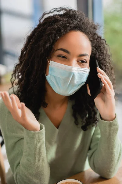 Mujer afroamericana rizada en máscara médica hablando en teléfono inteligente en la cafetería - foto de stock