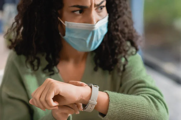 Verärgerte afrikanisch-amerikanische Frau in medizinischer Maske berührt Uhr am Handgelenk während des Wartens im Café — Stockfoto
