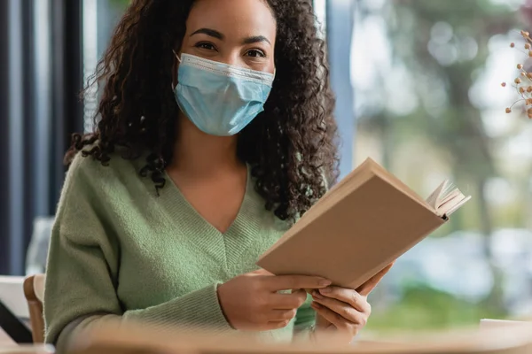 Mulher africana americana em máscara médica segurando livro no café — Fotografia de Stock