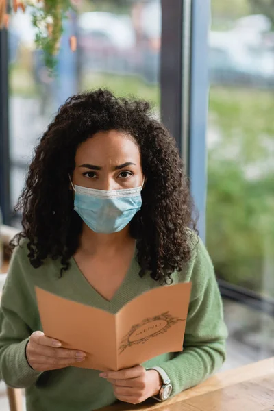 African american woman in medical mask holding menu and looking at camera — Stock Photo
