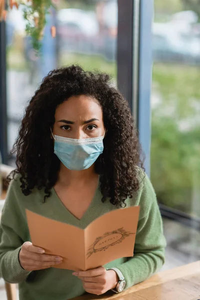 Africano americano mujer en médico máscara celebración menú y mirando cámara - foto de stock