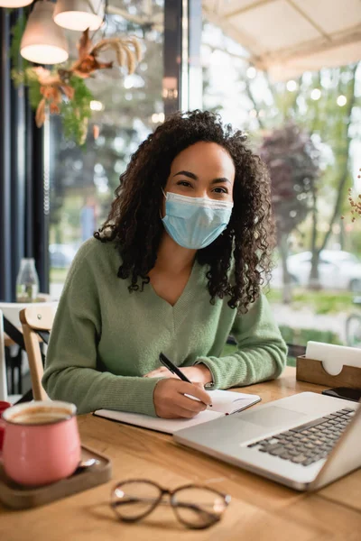 Mujer americana africana rizada en la escritura de la máscara médica en el cuaderno cerca del ordenador portátil en la cafetería - foto de stock