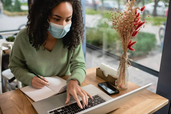 Афроамериканська жінка в медичній масці, використовуючи ноутбук біля смартфона і ноутбук на столі — стокове фото