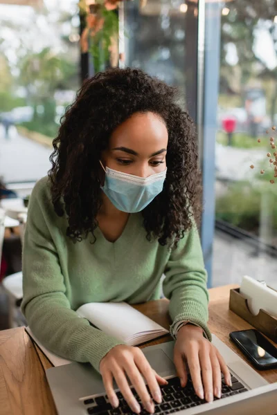 Афроамериканська жінка в медичній масці навчається онлайн, використовуючи ноутбук біля смартфона і зошит на столі. — стокове фото