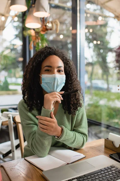 Afro-américaine en masque médical regardant la caméra près de gadgets et ordinateur portable sur la table dans un café — Photo de stock