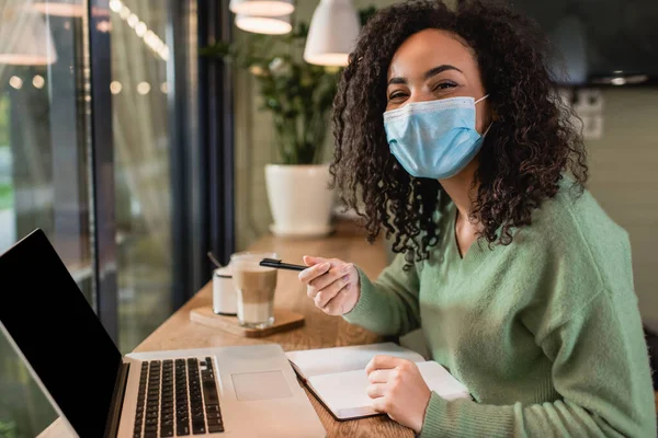 Mujer afroamericana en máscara médica que sostiene la pluma cerca del cuaderno y del ordenador portátil con la pantalla en blanco en la mesa - foto de stock