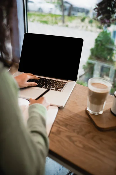 Partial view of african american woman typing on laptop with blank screen near glass of latte on table — Stock Photo