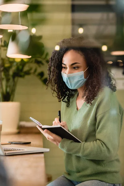 Mujer afroamericana en máscara médica que sostiene el cuaderno y la pluma mientras mira el portátil - foto de stock