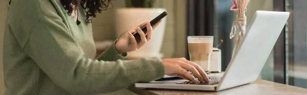 Vista parcial de la mujer afroamericana sosteniendo teléfono inteligente cerca de la computadora portátil y vidrio de café con leche en la cafetería, pancarta - foto de stock