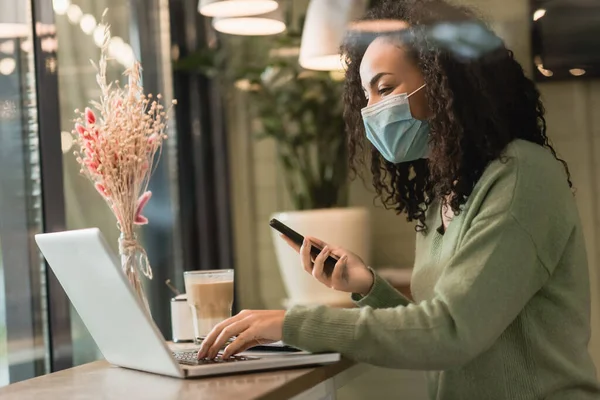 Freelancer americano africano encaracolado em máscara médica segurando smartphone perto de laptop e vidro de latte no café — Fotografia de Stock