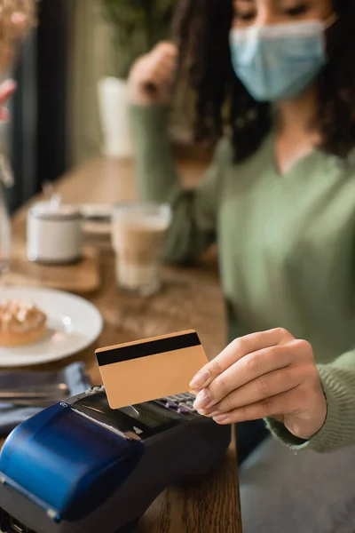 Tarjeta de crédito en mano de mujer afroamericana en máscara médica pagando en cafetería - foto de stock