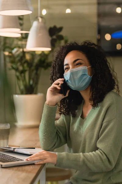 Африканська американка в медичній масці говорить на смартфоні біля ноутбука і зошиті на столі. — стокове фото