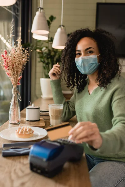 Mulher afro-americana em máscara médica pagando por torta e café no café — Fotografia de Stock