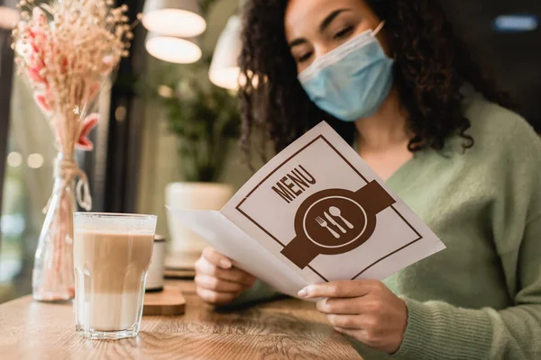 Mujer afroamericana en el menú de lectura máscara médica cerca de un vaso de café con leche en la cafetería - foto de stock