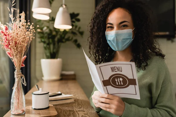 African american woman in medical mask holding brochure with menu lettering in cafe — Stock Photo