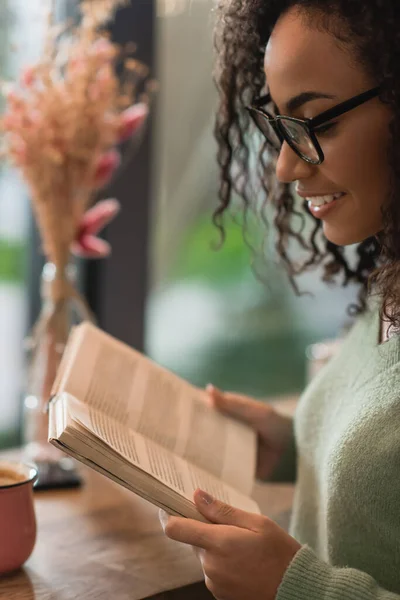 Mujer afroamericana feliz en anteojos libro de lectura en cafetería - foto de stock