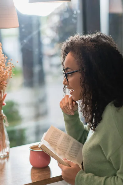 Vista laterale della donna afro-americana positiva negli occhiali da vista lettura libro in caffè — Foto stock