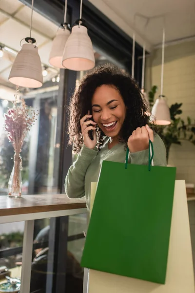 Cheerful african american woman talking on smartphone and holding shopping bags in cafe — Stock Photo