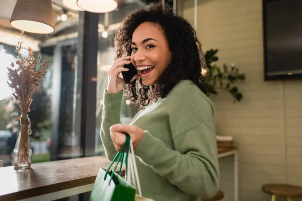 Sonriente mujer afroamericana hablando en el teléfono inteligente y sosteniendo bolsas de compras en la cafetería - foto de stock