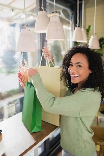 Mujer afroamericana feliz sosteniendo bolsas de compras en la cafetería - foto de stock