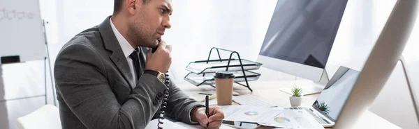 Comerciante serio hablando por teléfono mientras escribe en el cuaderno y mirando el ordenador portátil, bandera - foto de stock