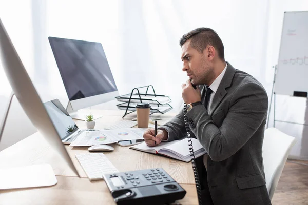 Serio commerciante che parla al telefono mentre scrive in notebook vicino a monitor e notebook, in primo piano sfocato — Foto stock