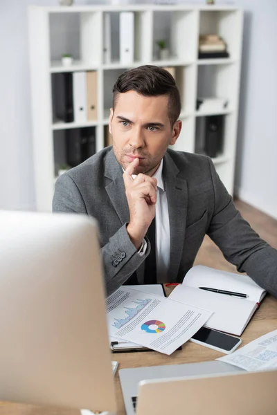 Comerciante reflexivo mirando monitor cerca de papeles con gráficos y teléfono inteligente con pantalla en blanco, primer plano borrosa - foto de stock