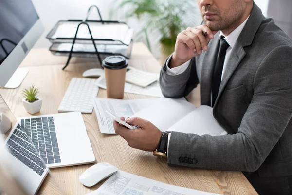 Ausgeschnittene Ansicht der Händlernachrichten auf dem Smartphone in der Nähe von Laptop, Papieren und Monitoren — Stockfoto