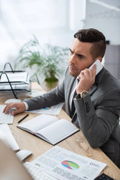 Serio uomo d'affari che parla su smartphone vicino a notebook e infografiche in ufficio, primo piano sfocato — Foto stock