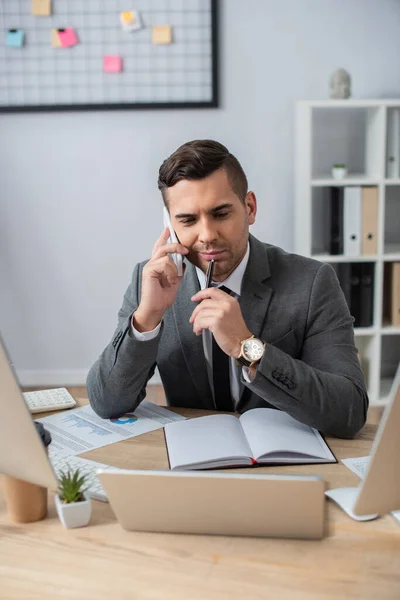 Lächelnder Geschäftsmann hält Stift in der Hand, während er auf dem Smartphone neben leerem Notizbuch und Laptop im verschwommenen Vordergrund spricht — Stockfoto