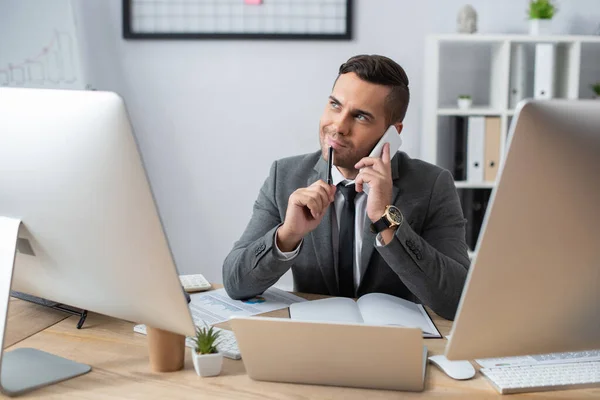 Comerciante sorrindo olhando para longe ao falar no telefone celular perto de laptop e monitores no local de trabalho — Fotografia de Stock