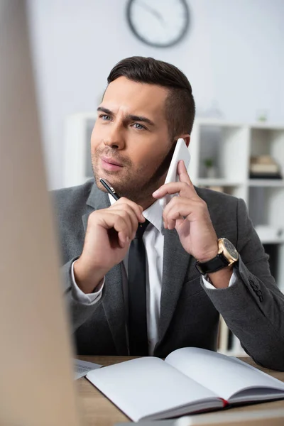 Thoughtful trader holding pen while talking on mobile phone in office, blurred foreground — Stock Photo
