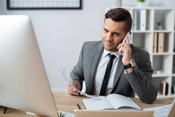 Sorridente, scettico commerciante parlando sul cellulare vicino notebook vuoto, primo piano sfocato — Foto stock