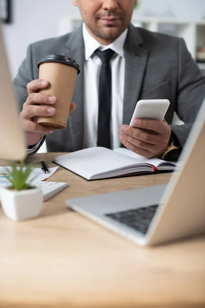 Обрізаний вид трейдера, який тримає каву, щоб піти під час обміну повідомленнями на смартфоні біля ноутбука на розмитому передньому плані — стокове фото