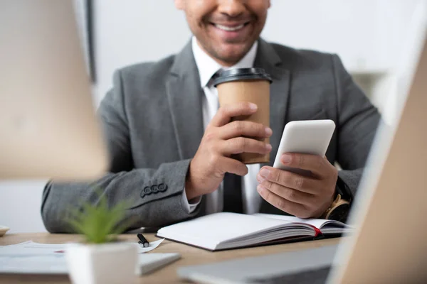 Vista cortada de homem de negócios sorridente conversando no smartphone enquanto segurando café para ir em primeiro plano desfocado — Fotografia de Stock