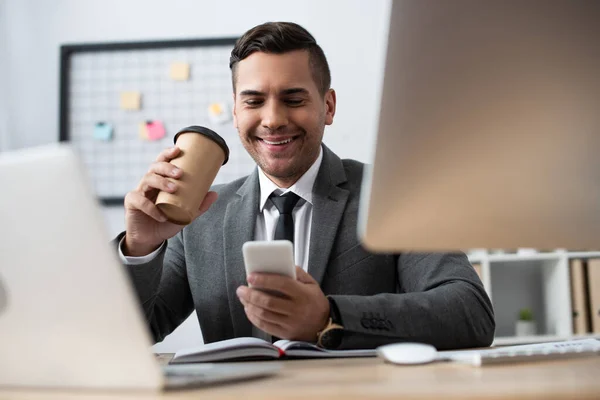 Trader sorridente che chiacchiera sullo smartphone mentre tiene il caffè in carica, in primo piano sfocato — Foto stock