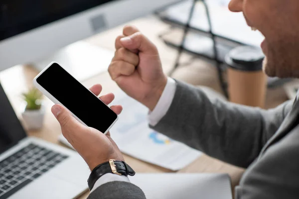 Vista recortada del comerciante emocionado mostrando gesto de victoria mientras sostiene el teléfono inteligente con pantalla en blanco, primer plano borrosa - foto de stock