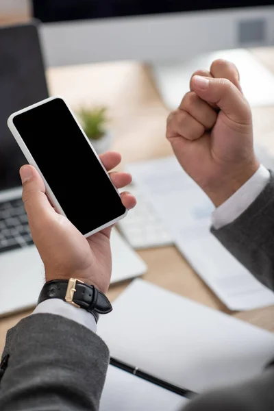 Vista parziale del trader di successo che mostra il gesto di vittoria mentre tiene il telefono cellulare con schermo vuoto, primo piano sfocato — Foto stock