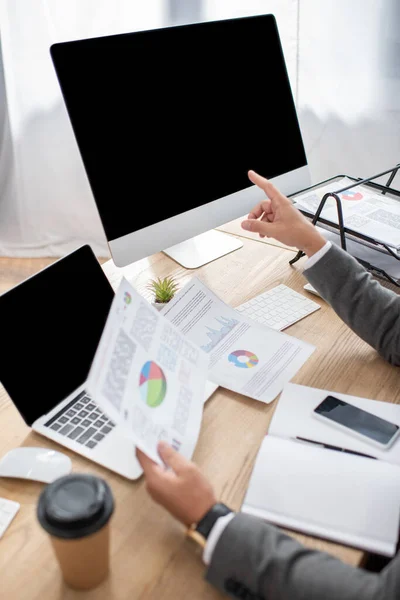 Cropped view of trader pointing with finger at monitor while holding infographics, blurred foreground — Stock Photo