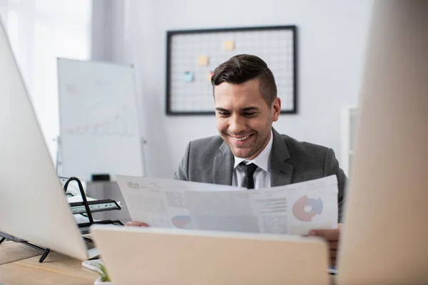 Comerciante feliz segurando infográficos perto de monitores em primeiro plano desfocado — Fotografia de Stock