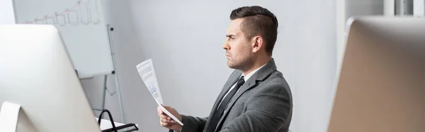 Seitenansicht des Händlers beim Betrachten des Dokuments, während er in der Nähe von Monitoren auf verschwommenem Vordergrund sitzt, Banner — Stockfoto