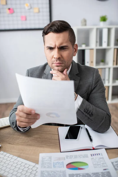 Comerciante reflexivo mirando documento cerca de portátil y teléfono inteligente con pantalla en blanco, primer plano borrosa - foto de stock