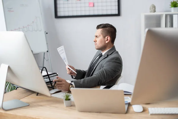 Seitenansicht des Geschäftsmannes beim Betrachten eines Dokuments in der Nähe von Laptop und Monitoren — Stockfoto