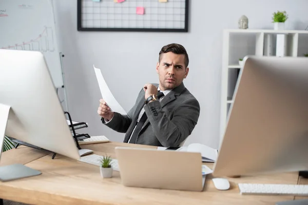 Uomo d'affari in possesso di carta mentre seduto sul posto di lavoro e guardando il monitor — Foto stock