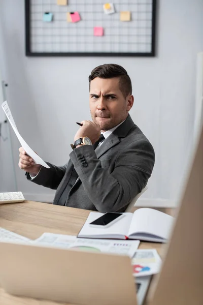 Nachdenklicher Händler hält Papier in der Nähe von Notizbuch, Smartphone und Laptop auf verschwommenem Vordergrund — Stockfoto