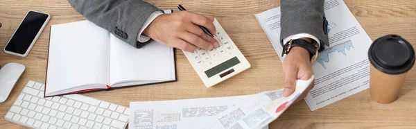 Vista parcial del comerciante que trabaja con la calculadora y las infografías cerca de portátil y café para llevar, bandera - foto de stock