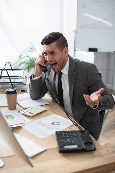 Comerciante irritado gritando enquanto falava no telefone fixo perto de papéis com infográficos, foreground borrado — Fotografia de Stock