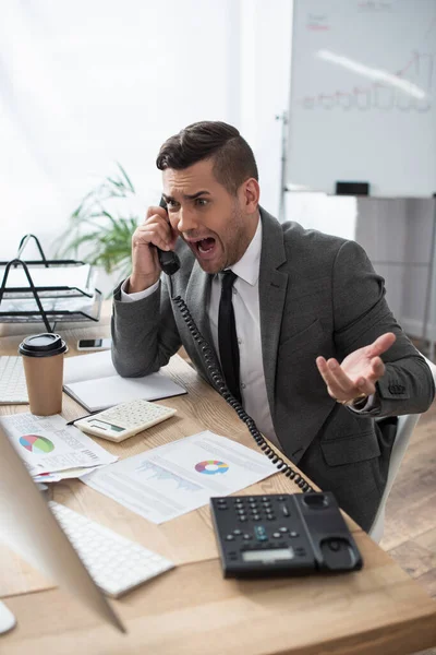 Comerciante enojado gritando mientras habla por teléfono fijo cerca de papeles con infografías, borrado primer plano - foto de stock