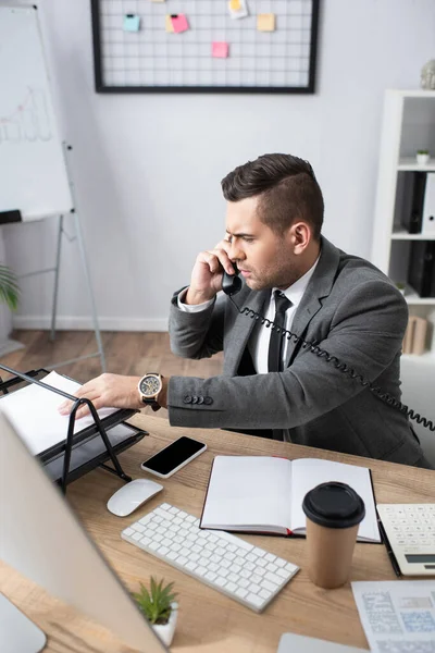 Seriöser Händler, der Papier nimmt, während er in der Nähe von Smartphone und Notebook telefoniert, verschwommener Vordergrund — Stockfoto